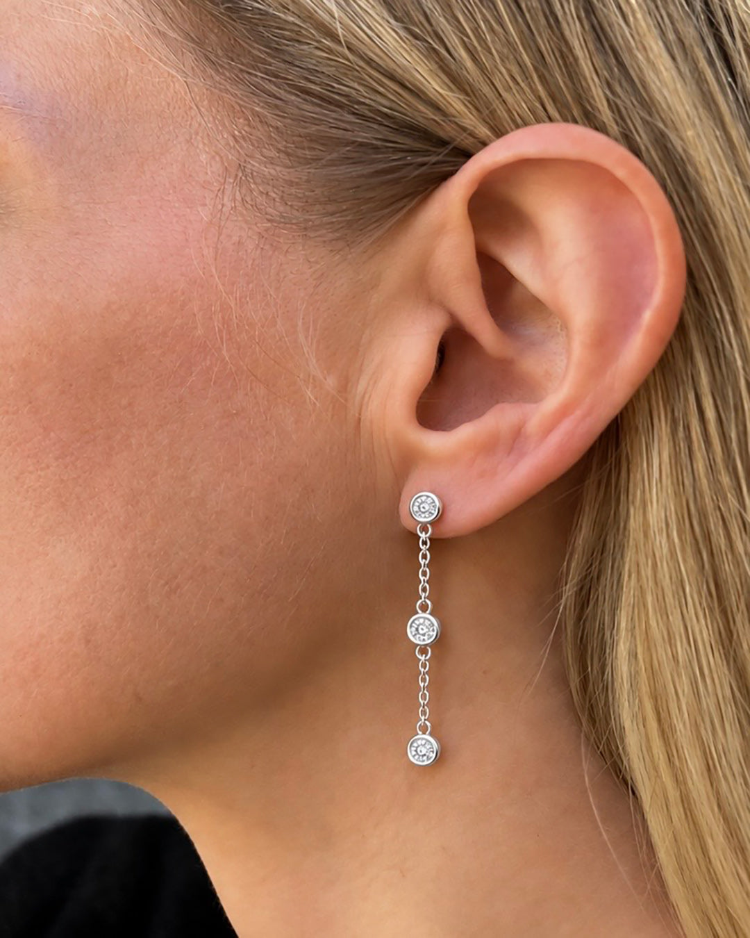 Close-up of a model wearing Bezel Set Diamond Drop Earrings in 14K white gold, featuring three round brilliant-cut diamonds per earring suspended along delicate cable chains.