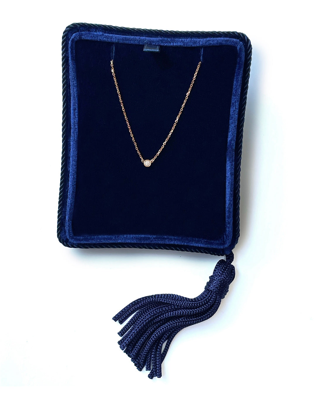 A delicate 14k rose gold chain necklace with a 0.05 carat round brilliant-cut diamond bezel set pendant, displayed inside a dark navy velvet jewelry box with a tassel.