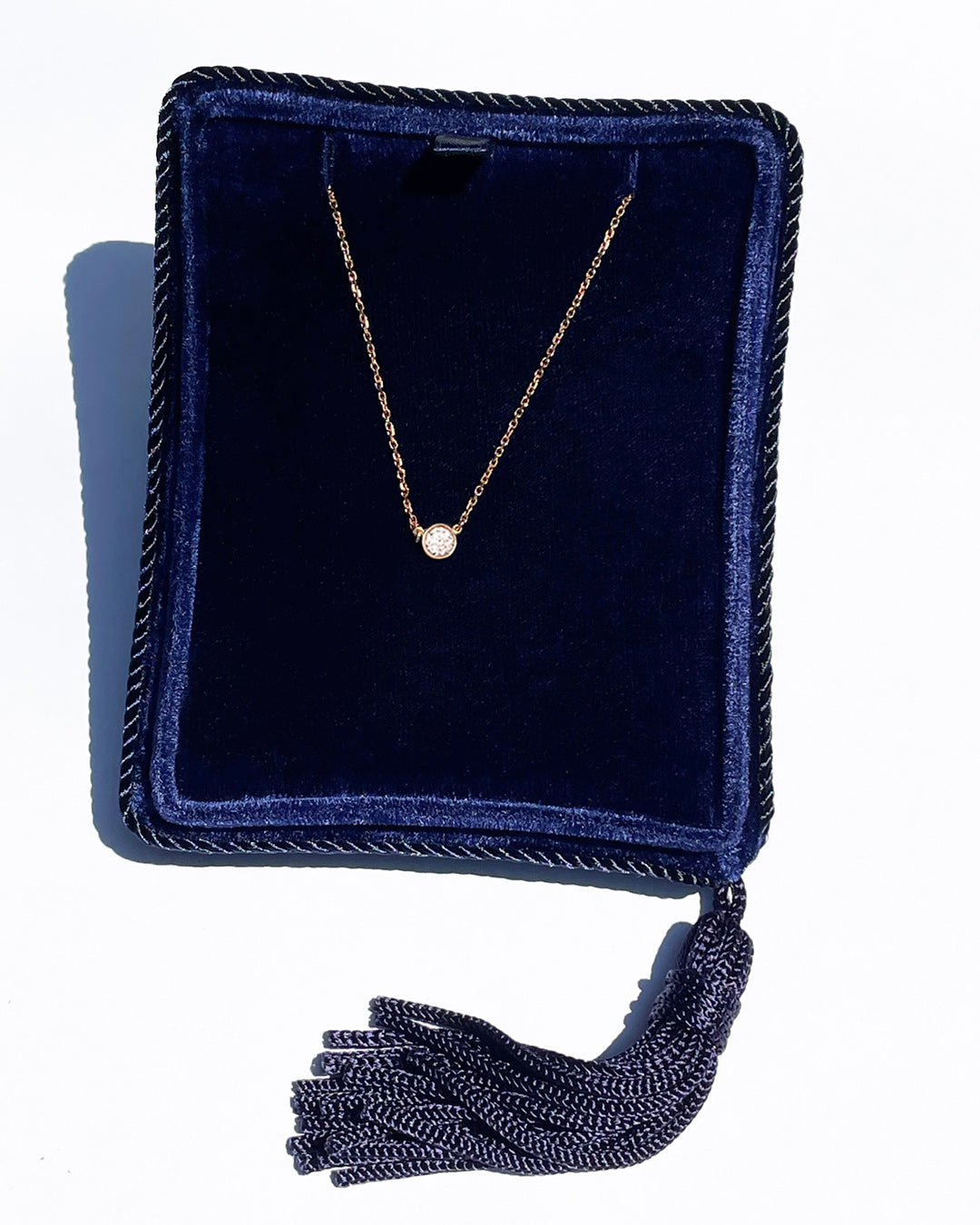 A delicate 14k rose gold chain necklace with a 0.15 carat round brilliant-cut diamond bezel set pendant, displayed inside a dark navy velvet jewelry box with a tassel.