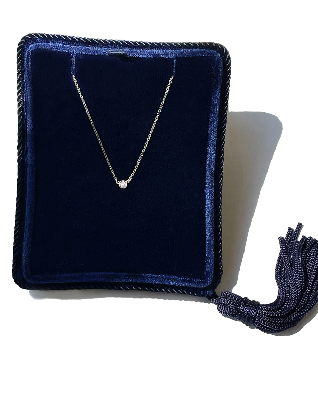 A delicate 14k white gold chain necklace with a 0.05 carat round brilliant-cut diamond bezel set pendant, displayed inside a dark navy velvet jewelry box with a tassel.