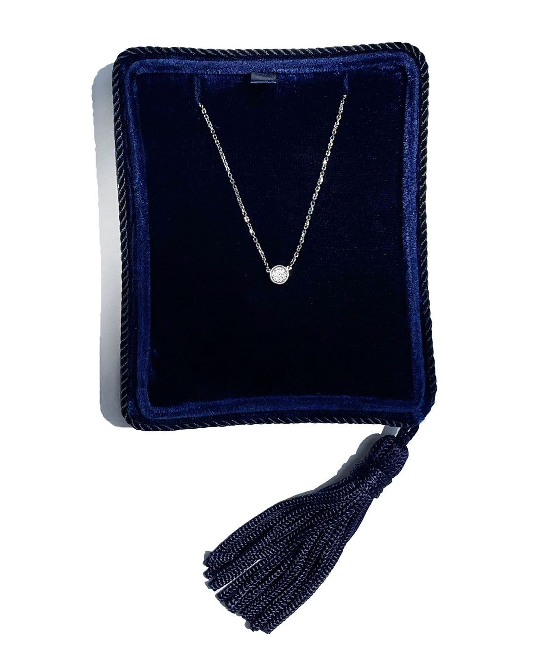 A delicate 14k white gold chain necklace with a 0.25 carat round brilliant-cut diamond bezel set pendant, displayed inside a dark navy velvet jewelry box with a tassel.