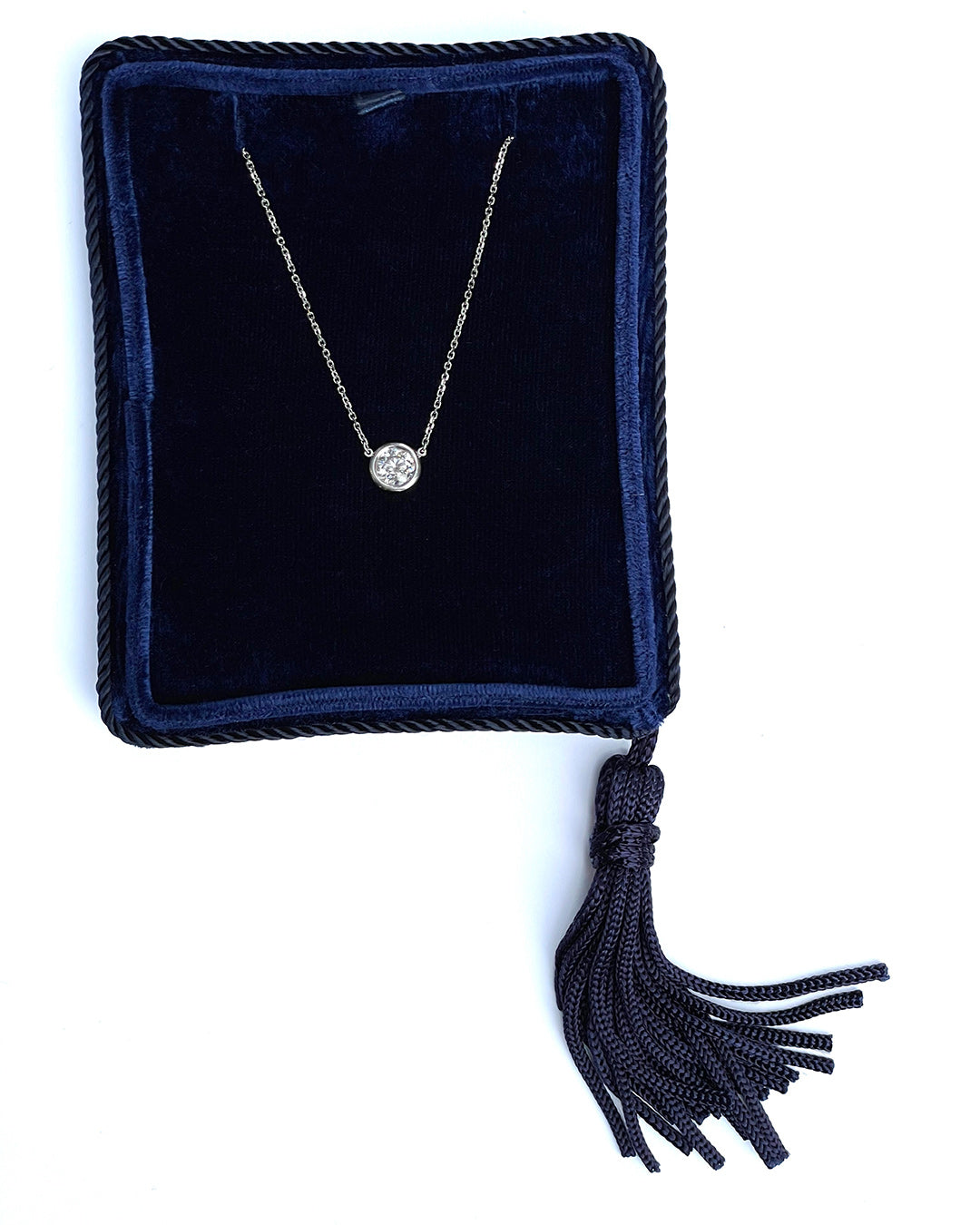 A delicate 14k white gold chain necklace with a 0.75 carat round brilliant-cut diamond bezel set pendant, displayed inside a dark navy velvet jewelry box with a tassel.