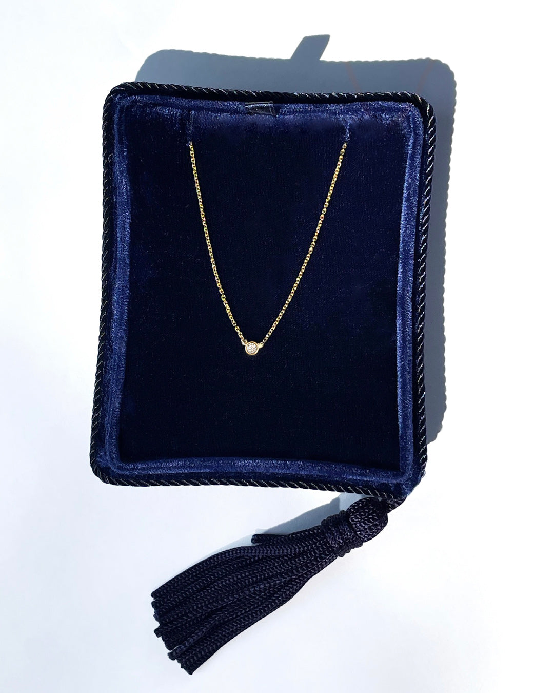 A delicate 14k yellow gold chain necklace with a 0.05 carat round brilliant-cut diamond bezel set pendant, displayed inside a dark navy velvet jewelry box with a tassel.