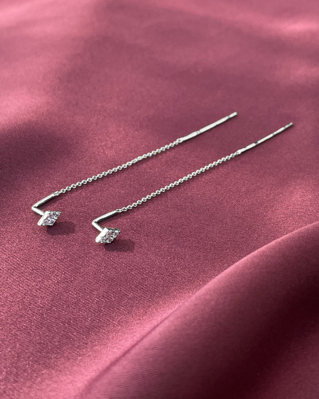A pair of diamond cluster threader earrings in 14k white gold displayed on a draped dusty rose satin fabric, showing the fine gold chains, polished threader bars,and diamond cluster details.