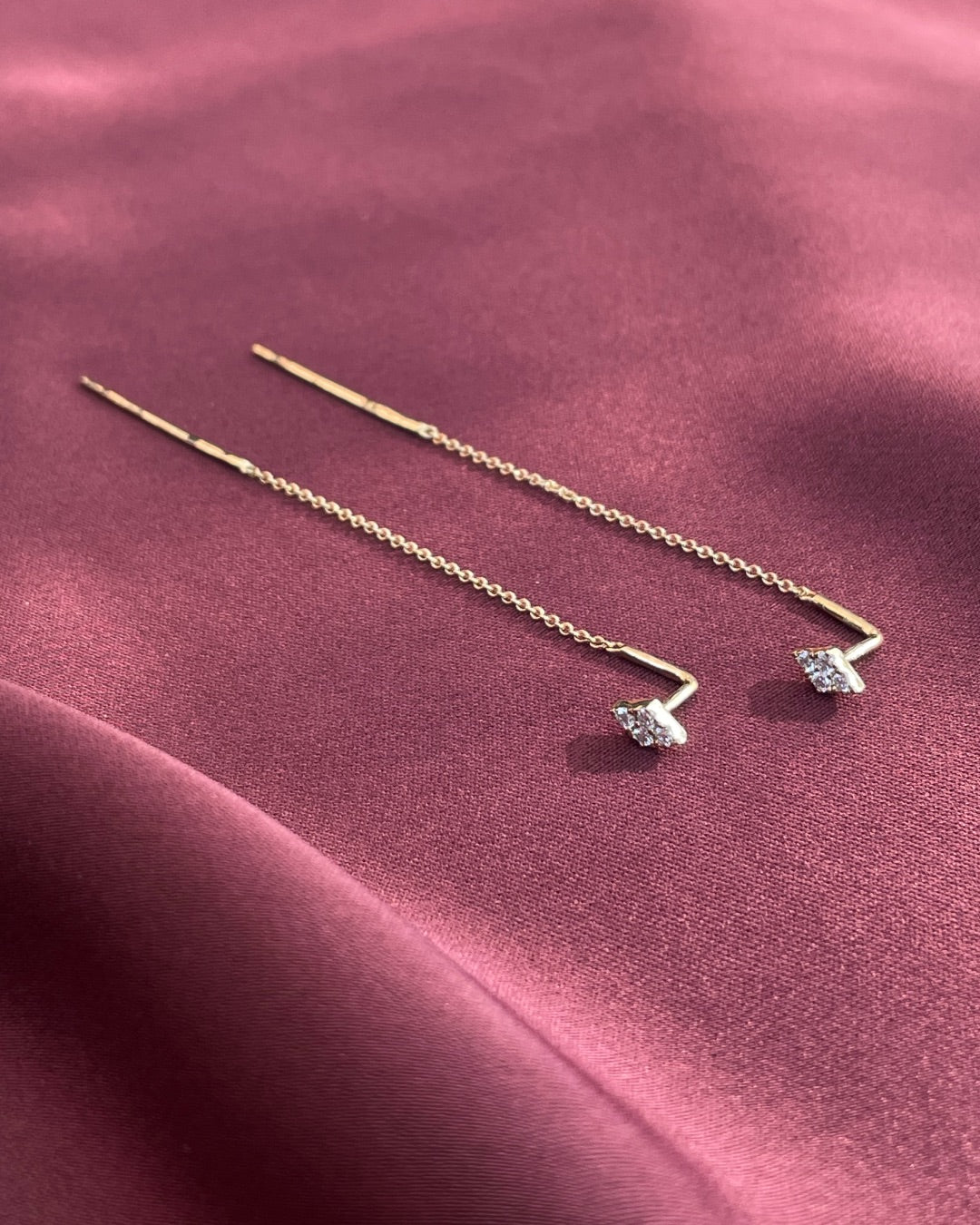 A pair of diamond cluster threader earrings in 14k yellow gold displayed on a draped dusty rose satin fabric, showing the fine gold chains, polished threader bars,and diamond cluster details.