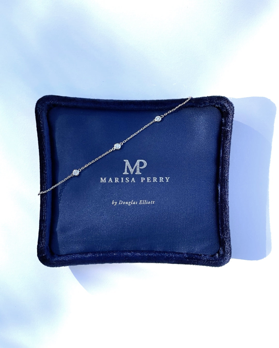White gold diamond station bracelet displayed across a navy Marisa Perry jewelry box, featuring bezel-set round diamonds.