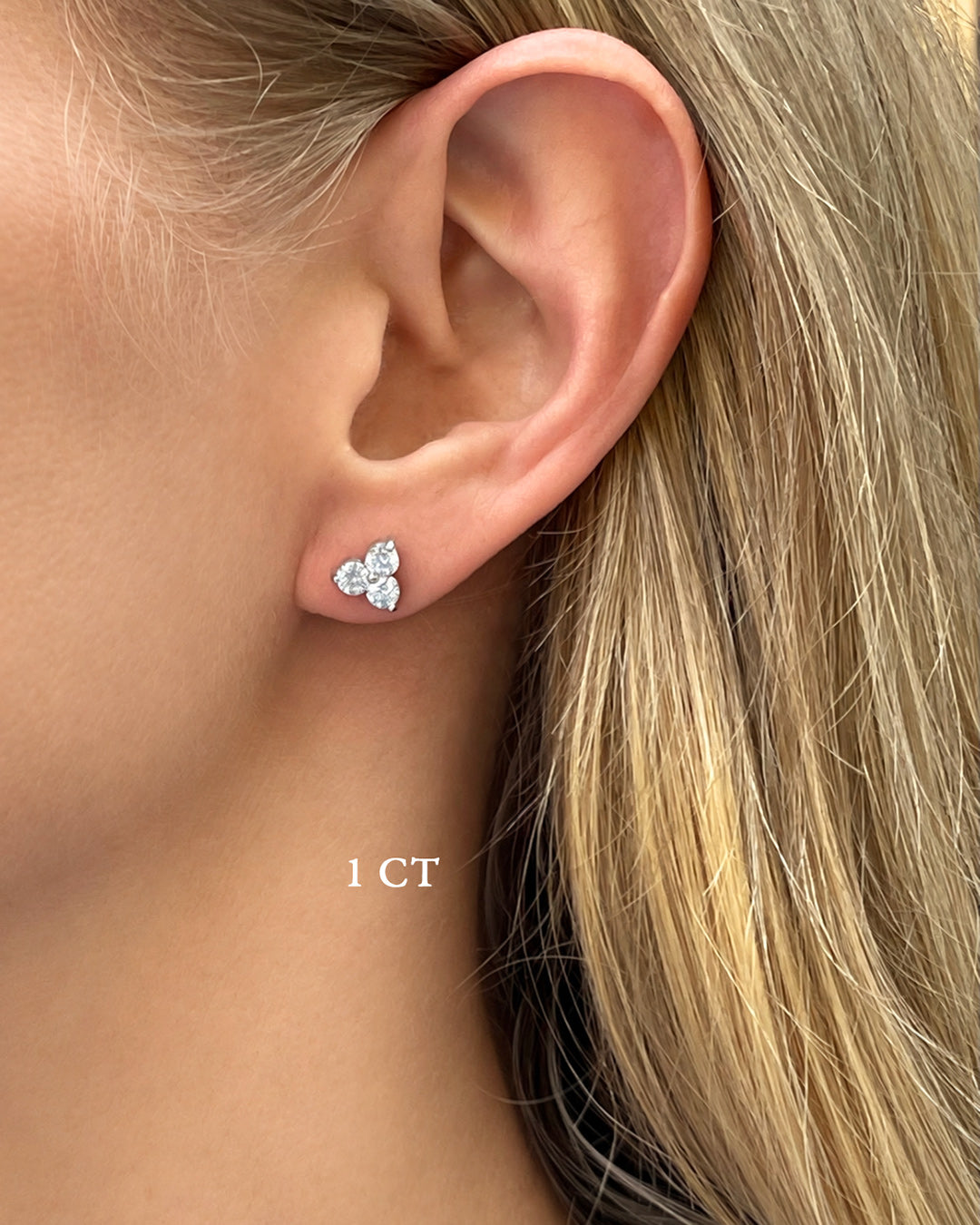 A blonde model wearing large diamond trilogy stud earrings in 14k white gold with a total of 1 carat of round brilliant-cut diamonds.
