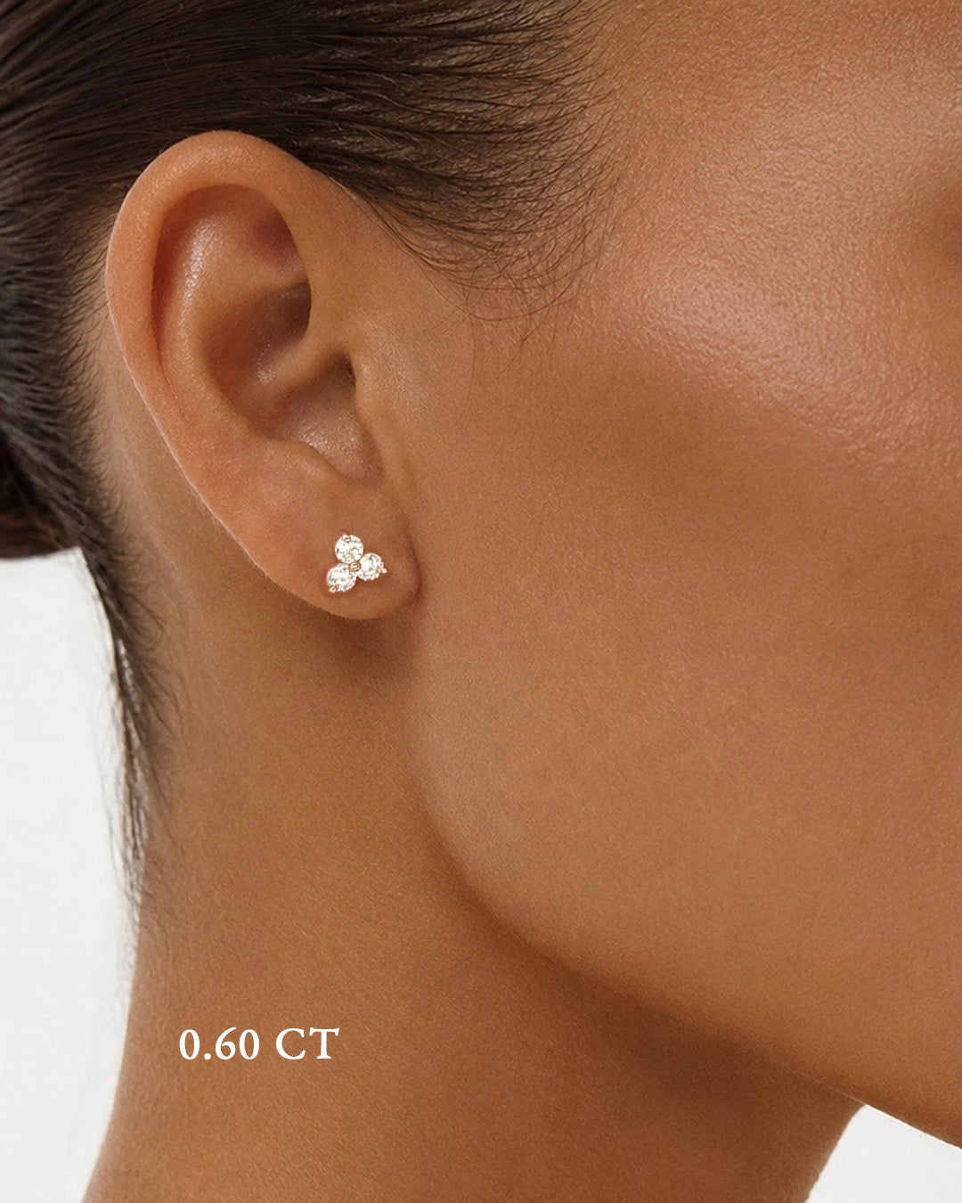 A close-up of a model wearing medium diamond trilogy stud earrings in 14K rose gold featuring 0.60 TCW of round brilliant-cut diamonds.