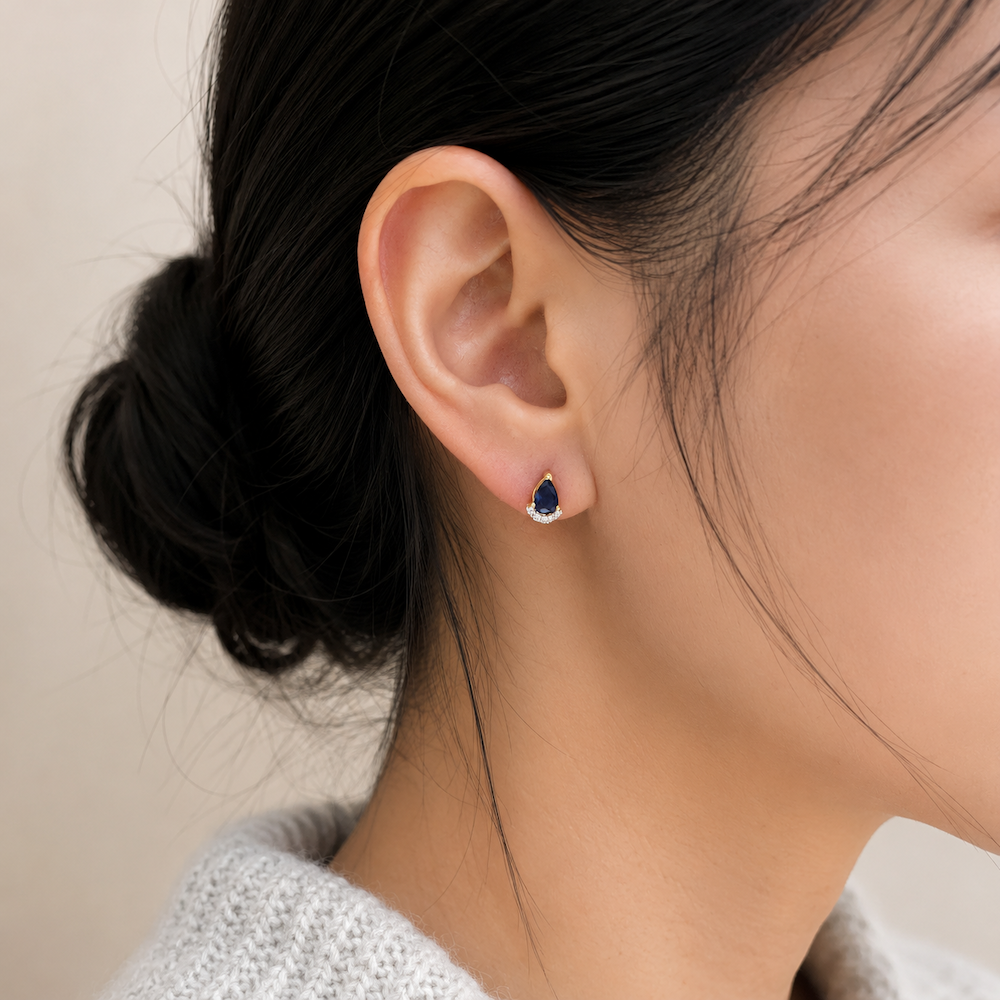 Close-up of a model’s ear wearing a pear-shaped blur sapphire and diamond stud earring with a blurred background.