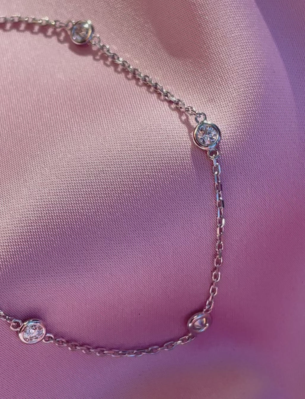 Bezel Set Diamond Station Bracelet in 14K white gold displayed on pink fabric, highlighting the round diamonds, delicate chain and lobster clasp.