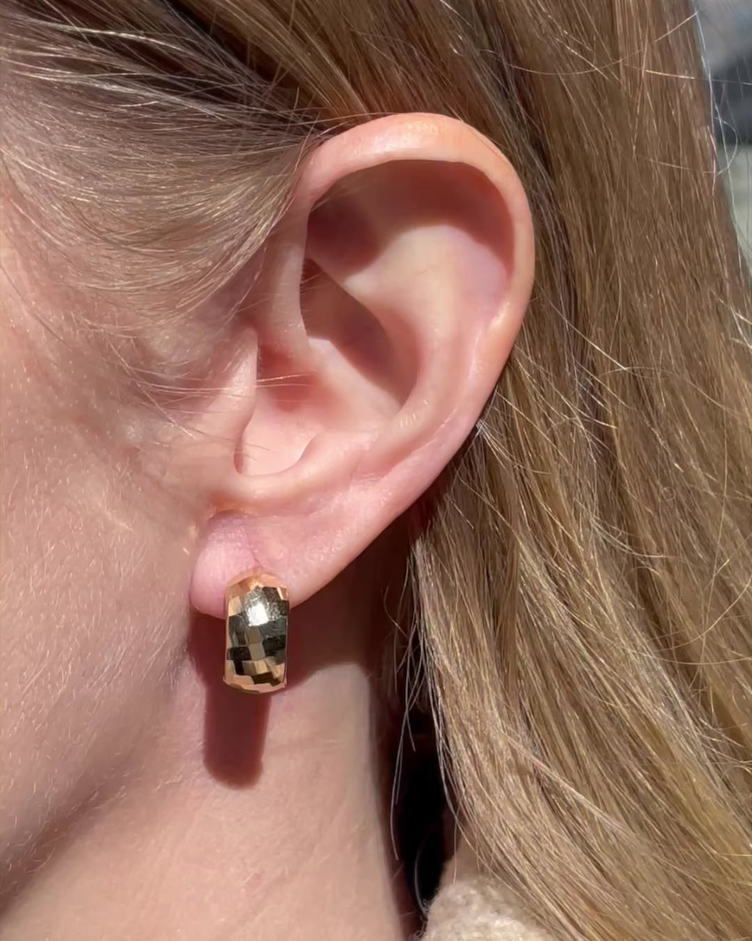 Close-up of a model wearing textured wide huggie hoop earrings, moving gently from side to side as the earring glistens in the sunlight.
