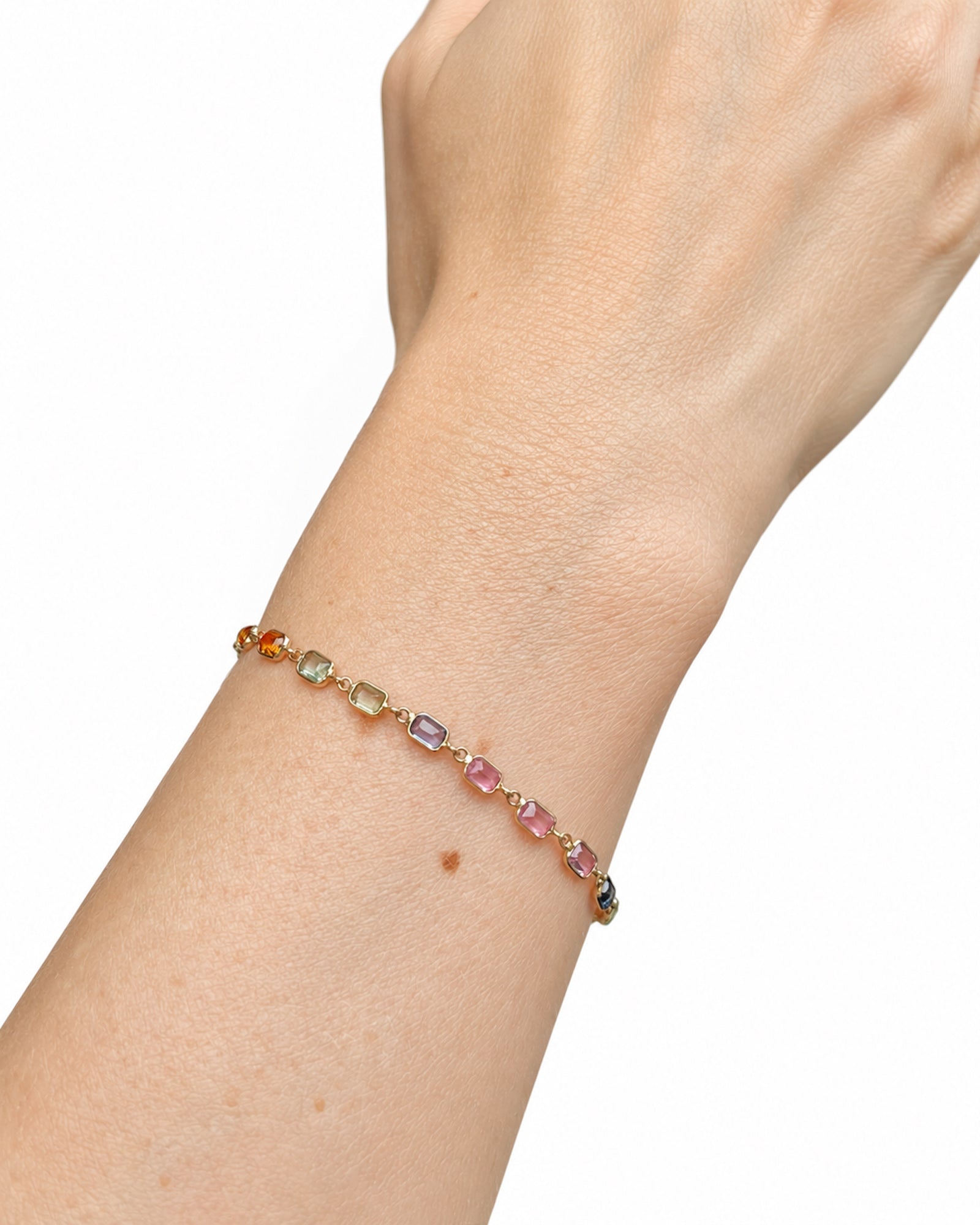 Close-up of a wrist wearing a Rainbow Sapphire Bezel Set Tennis Bracelet featuring emerald-cut natural multicolor sapphires set in 14K yellow gold.