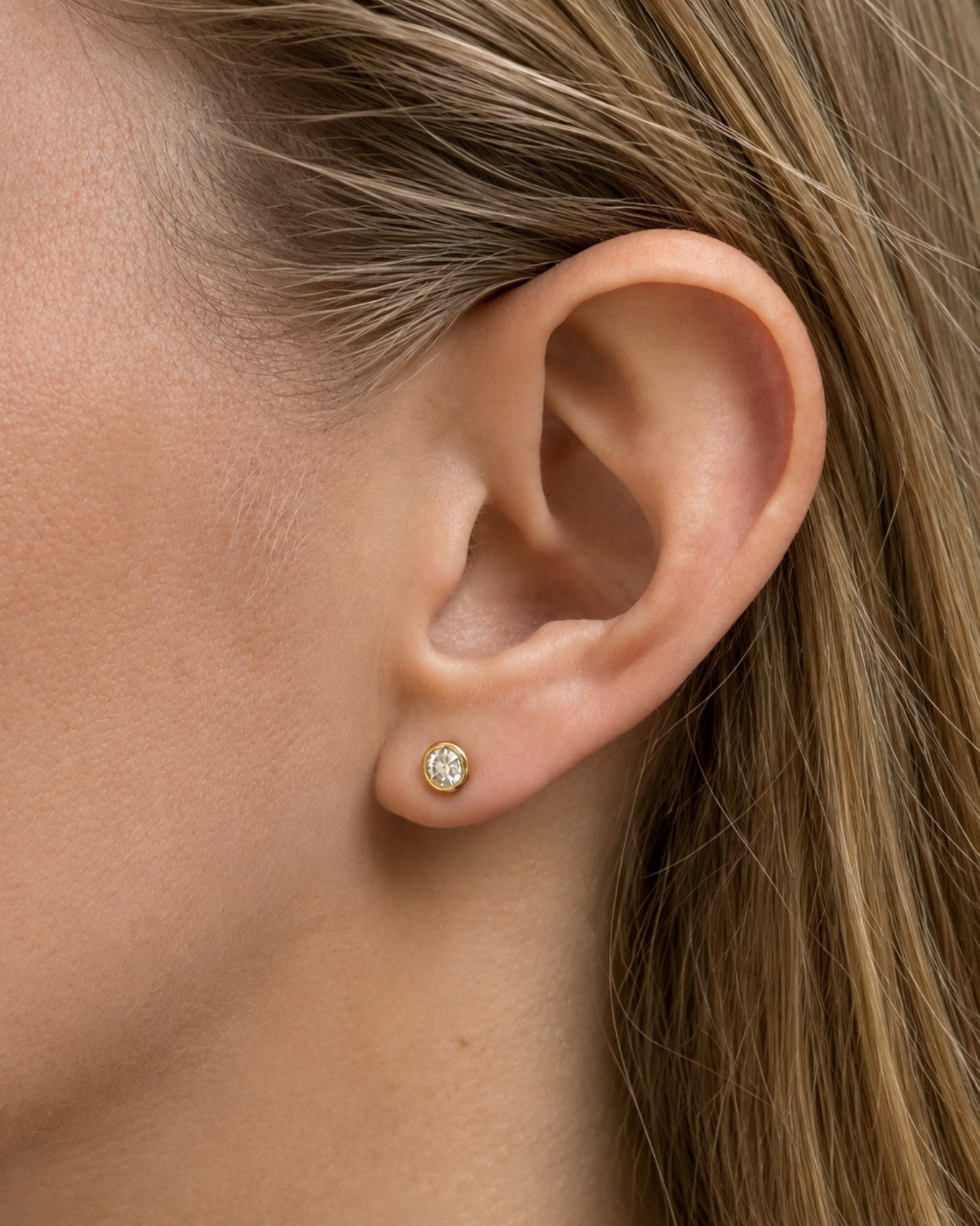 A close-up of a model wearing round brilliant bezel set diamond stud earrings. 